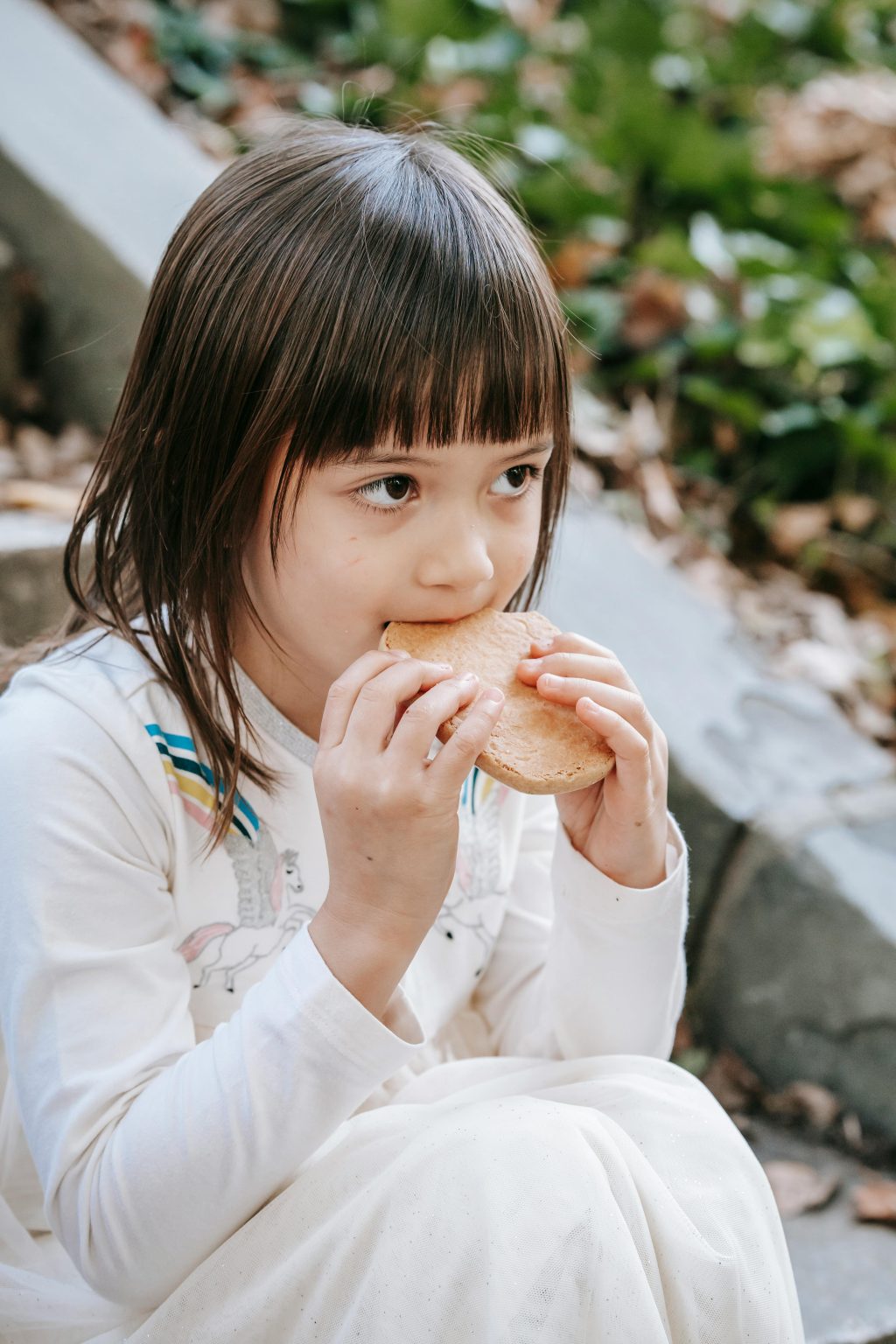 Cute little girl in white dress sitting on step and biting tasty cookie